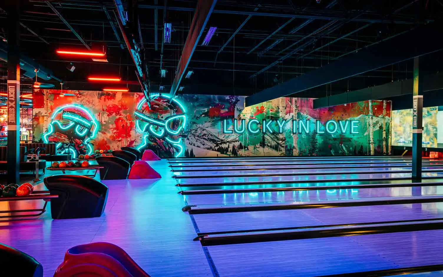 Colorful bowling balls on return rack with bowling center and neon lights in the background.