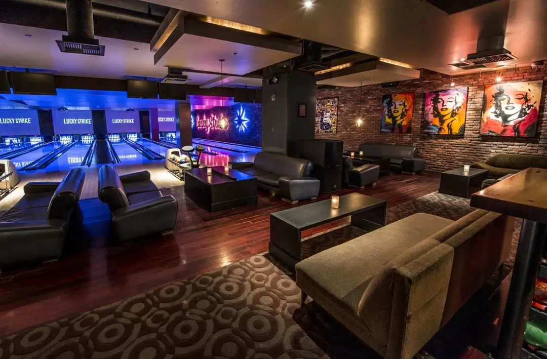 Upscale Lucky Strike bowling lounge with bookshelves, plush seating, high-top tables, and glowing lanes under blue lighting.