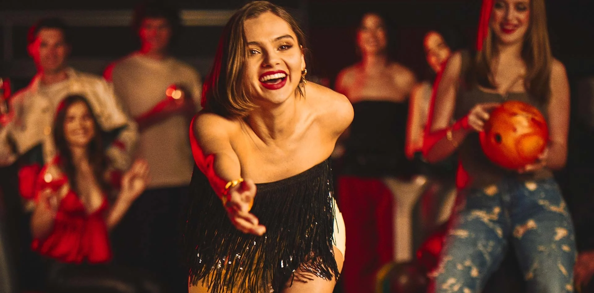 Woman in a fringed top releases a red bowling ball down the lane, smiling as friends cheer behind her in a warmly lit bowling lounge