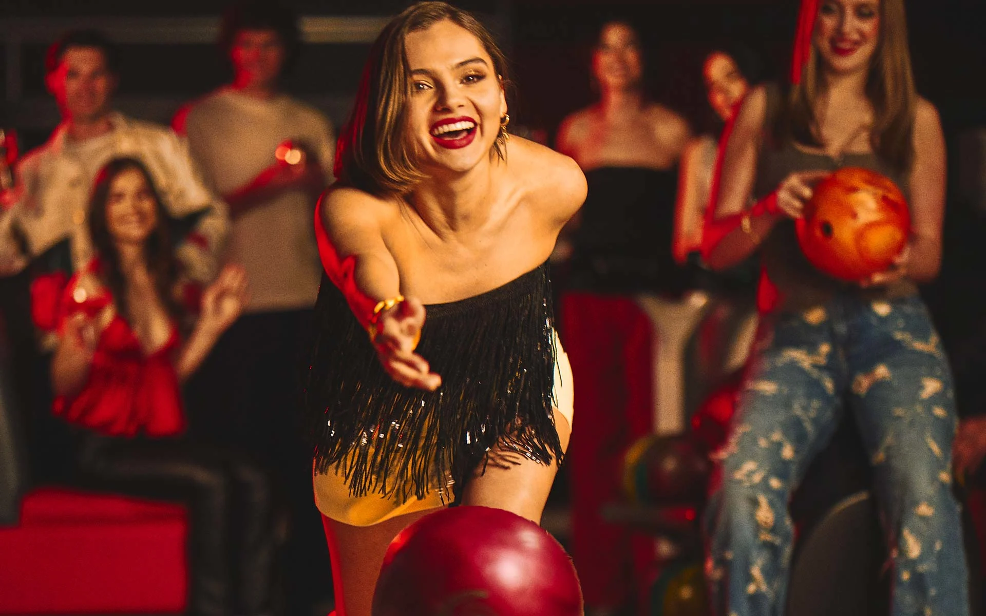 Woman in a fringed top releases a red bowling ball down the lane, smiling as friends cheer behind her in a warmly lit bowling lounge