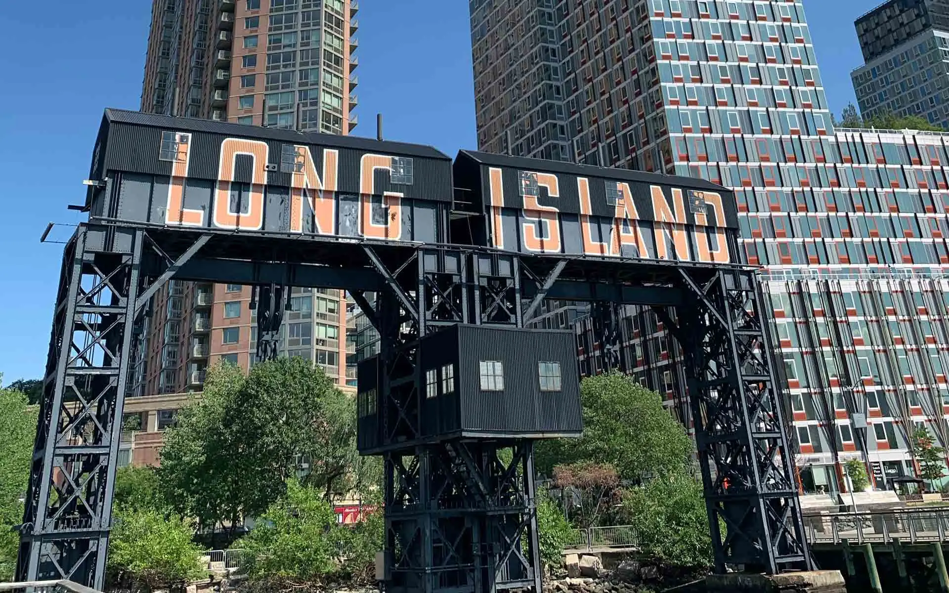 Long Island City sign on a black gantry with high-rise buildings and greenery along the Queens waterfront.