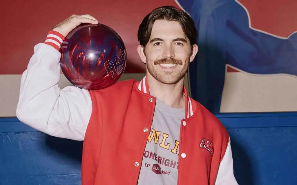 Smiling man in AMF varsity jacket balancing a bowling ball on his shoulder