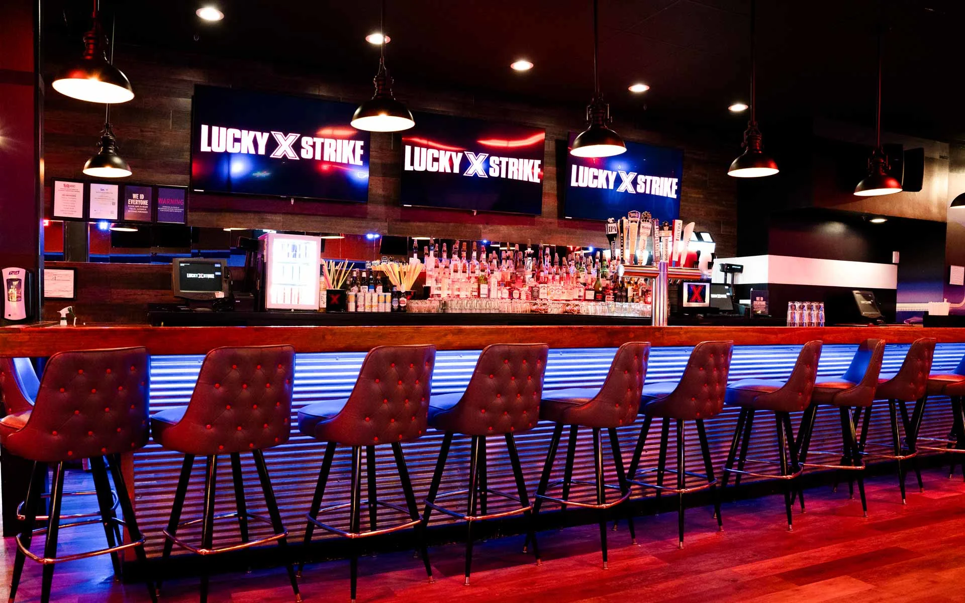 Upscale bar at Lucky Strike bowling alley with leather bar stools, glowing blue underlighting, beer taps, and large digital screens displaying the Lucky Strike logo.