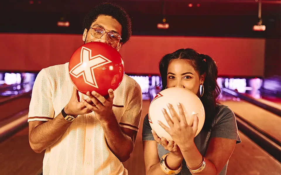 Two people holding bowling balls with bowling lanes in the background