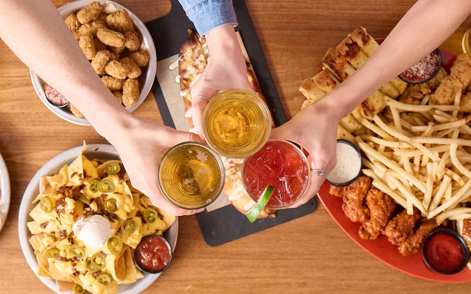 Top-down view of friends toasting drinks over a table filled with shareable food, including fries, nachos, chicken tenders, pretzels, and beer at a bowling center.