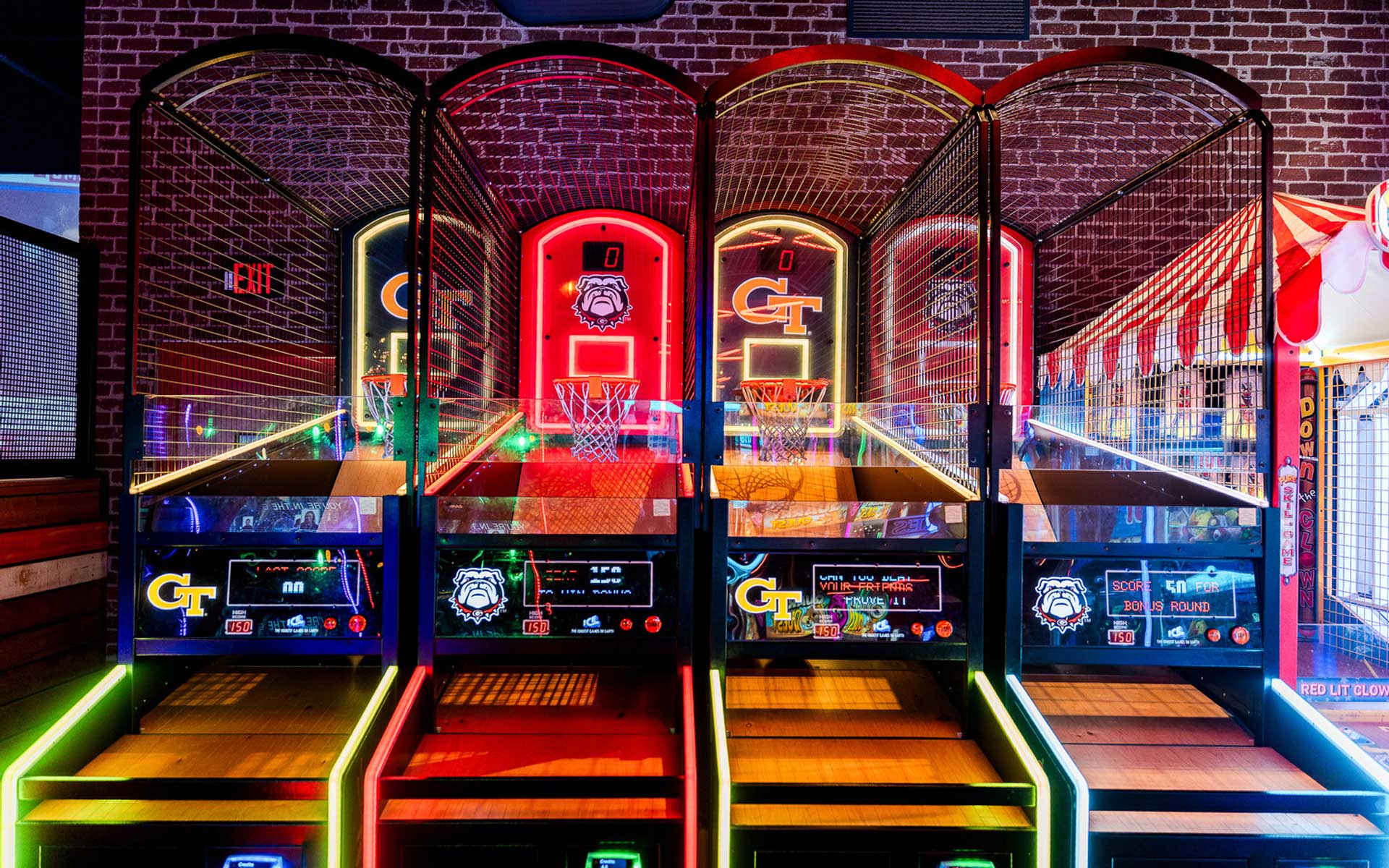 Row of illuminated basketball arcade games set against a brick wall, with glowing scoreboards, team logos, and bright neon accents creating a competitive, game-day atmosphere.