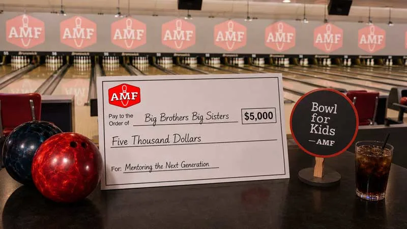 Diverse group celebrates an AMF Bowling fundraiser with donation check, Bowl for Kids paddle, drinks, and bowling balls.