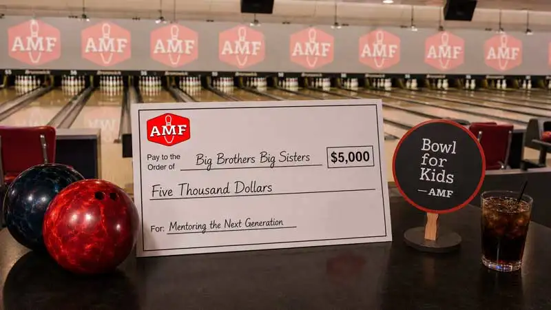 Diverse group celebrates an AMF Bowling fundraiser with donation check, Bowl for Kids paddle, drinks, and bowling balls.