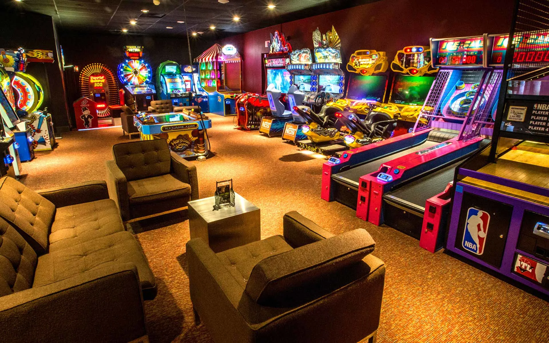 Spacious arcade area featuring a mix of classic and modern games, including racing simulators, skee-ball lanes, and prize machines, with cozy lounge seating and warm lighting for a relaxed, social atmosphere.