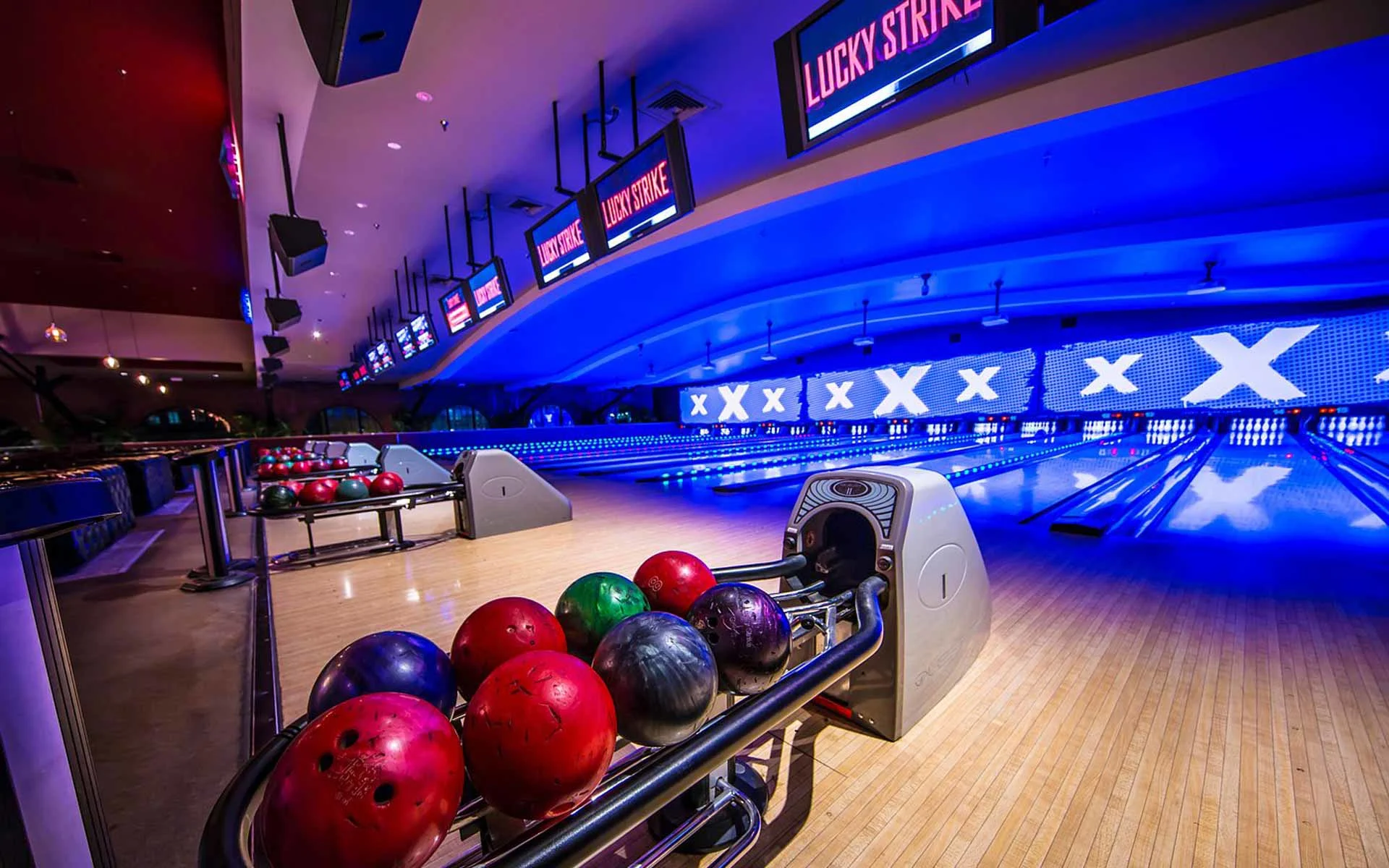 Neon-lit Lucky Strike bowling lanes with ball returns, overhead scoring monitors, and glowing lane displays in a high-energy environment.