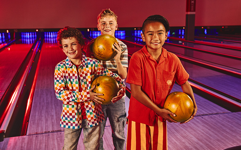 Three boys with bowling balls standing in front of bowling lanes.