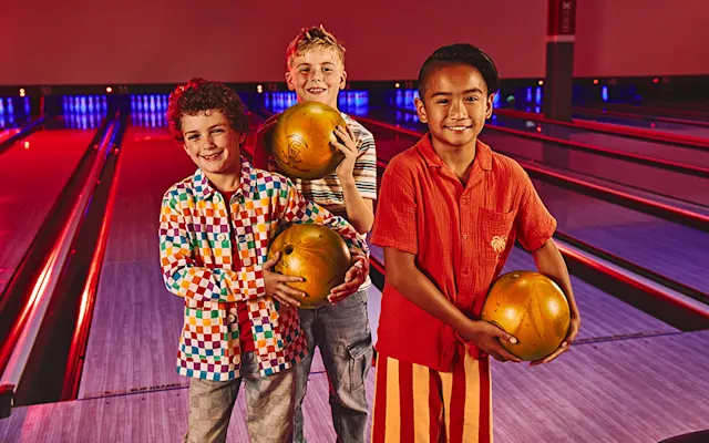 Three boys with bowling balls standing in front of bowling lanes.