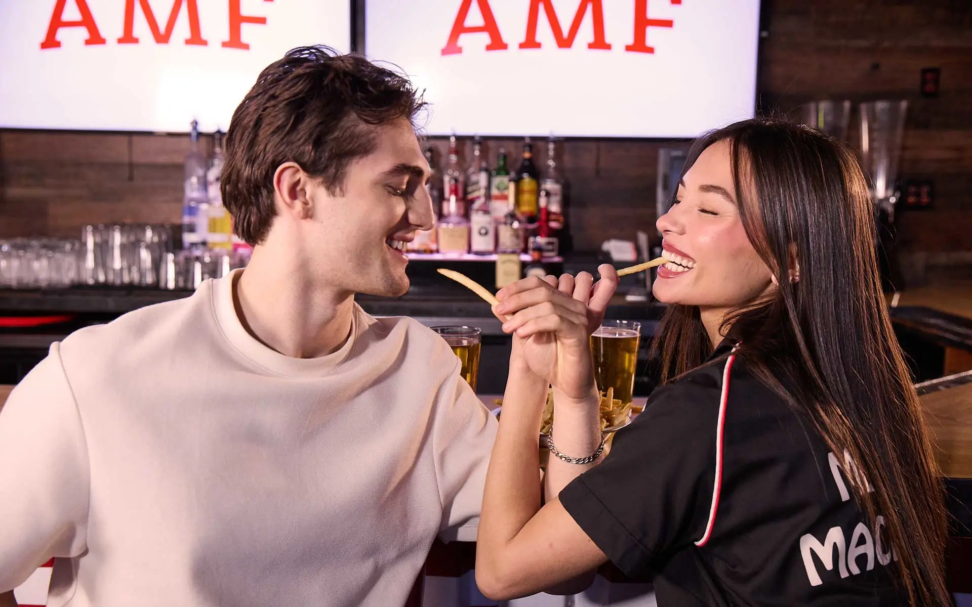 Couple enjoying drinks and sharing fries at a lively AMF bowling center bar, smiling and interacting in a casual, social date-night setting.