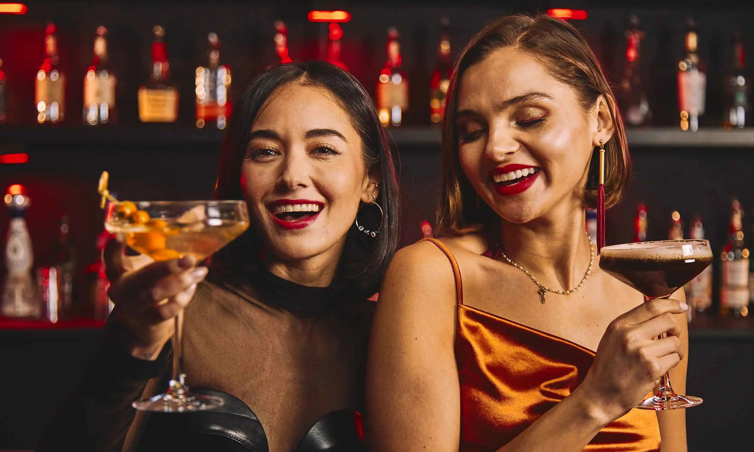 Two women smiling and toasting cocktails at a bar inside a bowling entertainment venue.