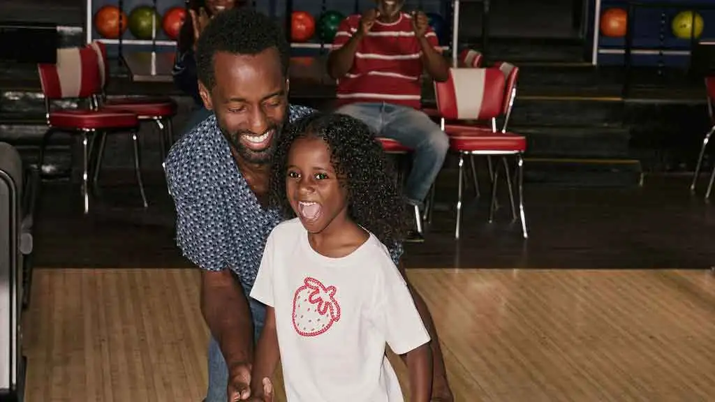 A father and daughter standing on a bowling lane approach as the father teaches the daughter how to throw a bowling ball.