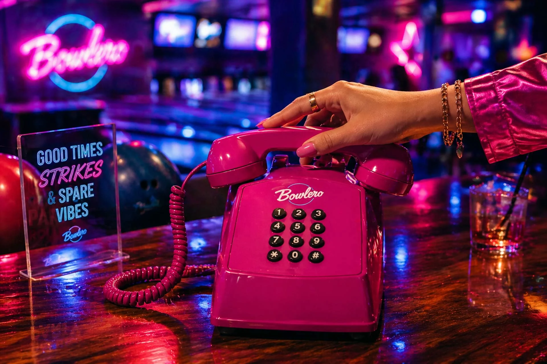 Hand placing a neon magenta retro phone on a glossy bowling lane table inside a vibrant Bowlero with colorful lights and lanes in the background.