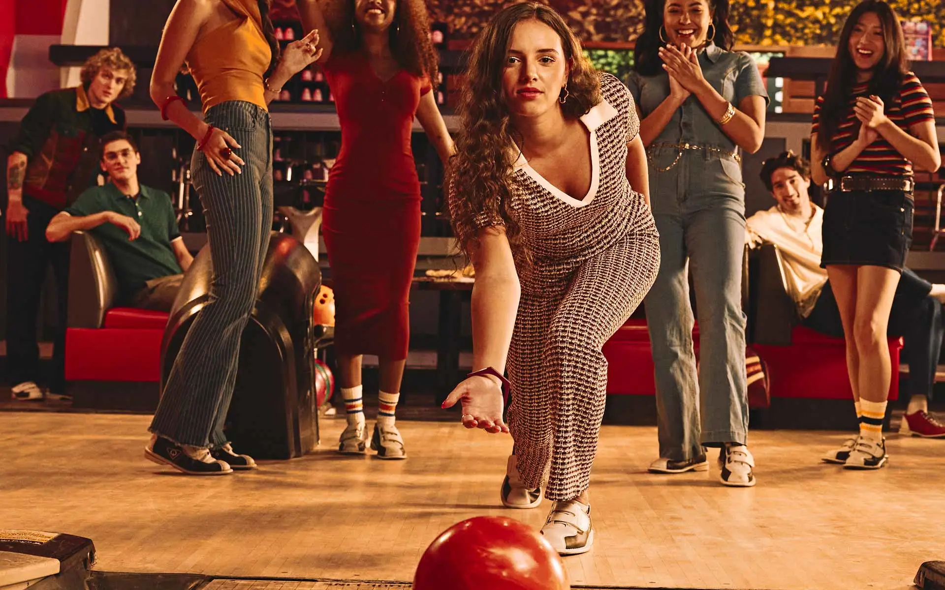 A group of friends cheer as a woman rolls a red bowling ball down the lane at Lucky Strike, captured mid-throw in a lively, neon-lit bowling alley.