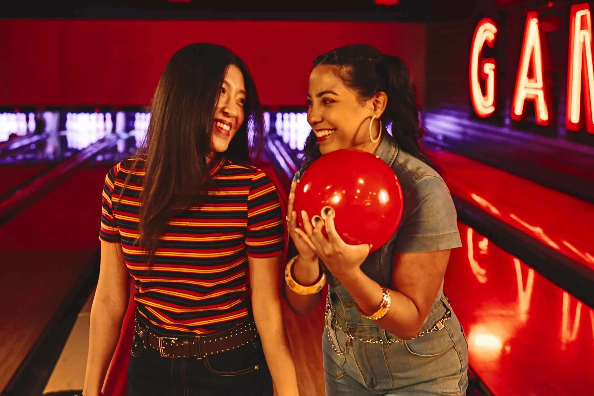Two friends smiling and standing at a bowling lane, with one holding a red bowling ball.