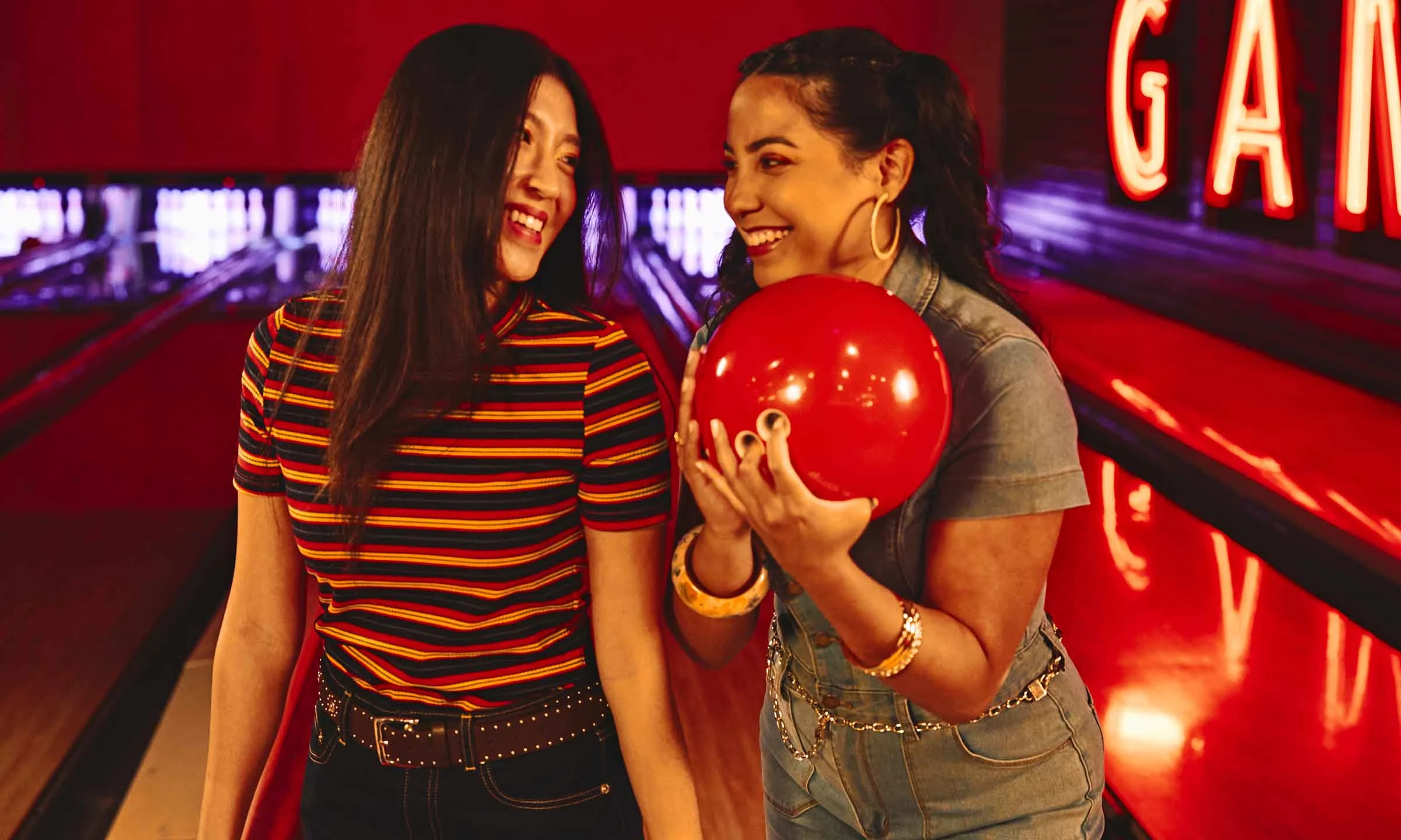 Two friends smiling and standing at a bowling lane, with one holding a red bowling ball.