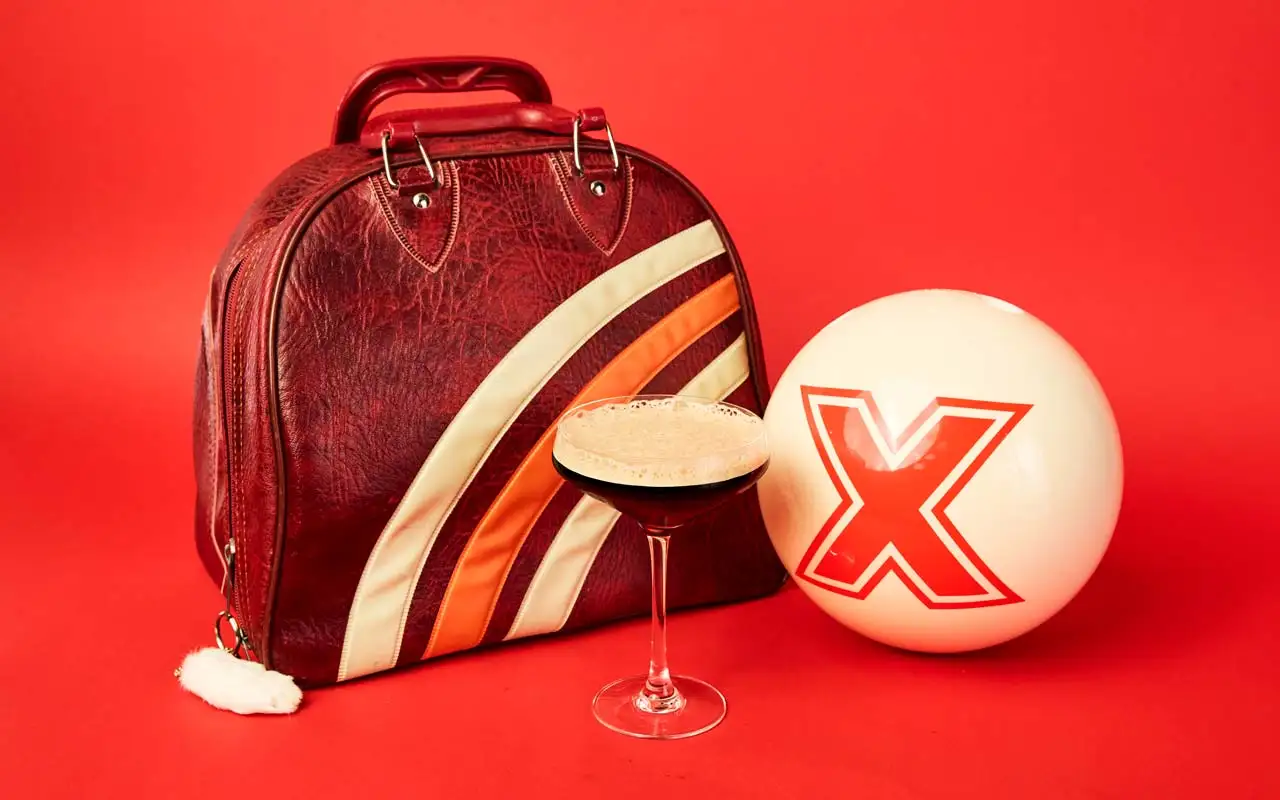 Vintage bowling bag, espresso martini, and a white bowling ball with a red X logo arranged against a bold red backdrop, blending retro bowling style with elevated cocktails.