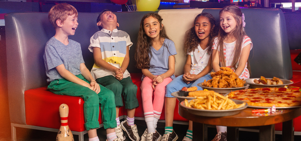Four kids laugh together at a birthday party booth in a bowling alley while pizza, fries, and chicken tenders sit on the table and confetti falls around them.