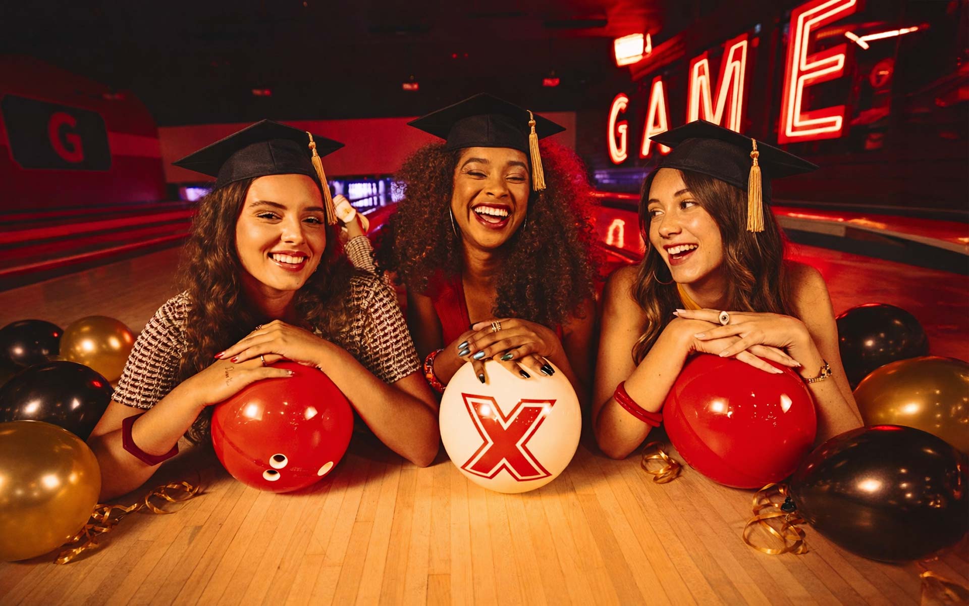 Three young women smile and lean on bowling balls at the front of a polished lane inside a modern, neon-lit bowling alley, with a sleek black graduation cap and gold tassel resting in the foreground and a small “Congrats Grad” card placed subtly on the wood surface, all integrated with warm red and orange ambient lighting.