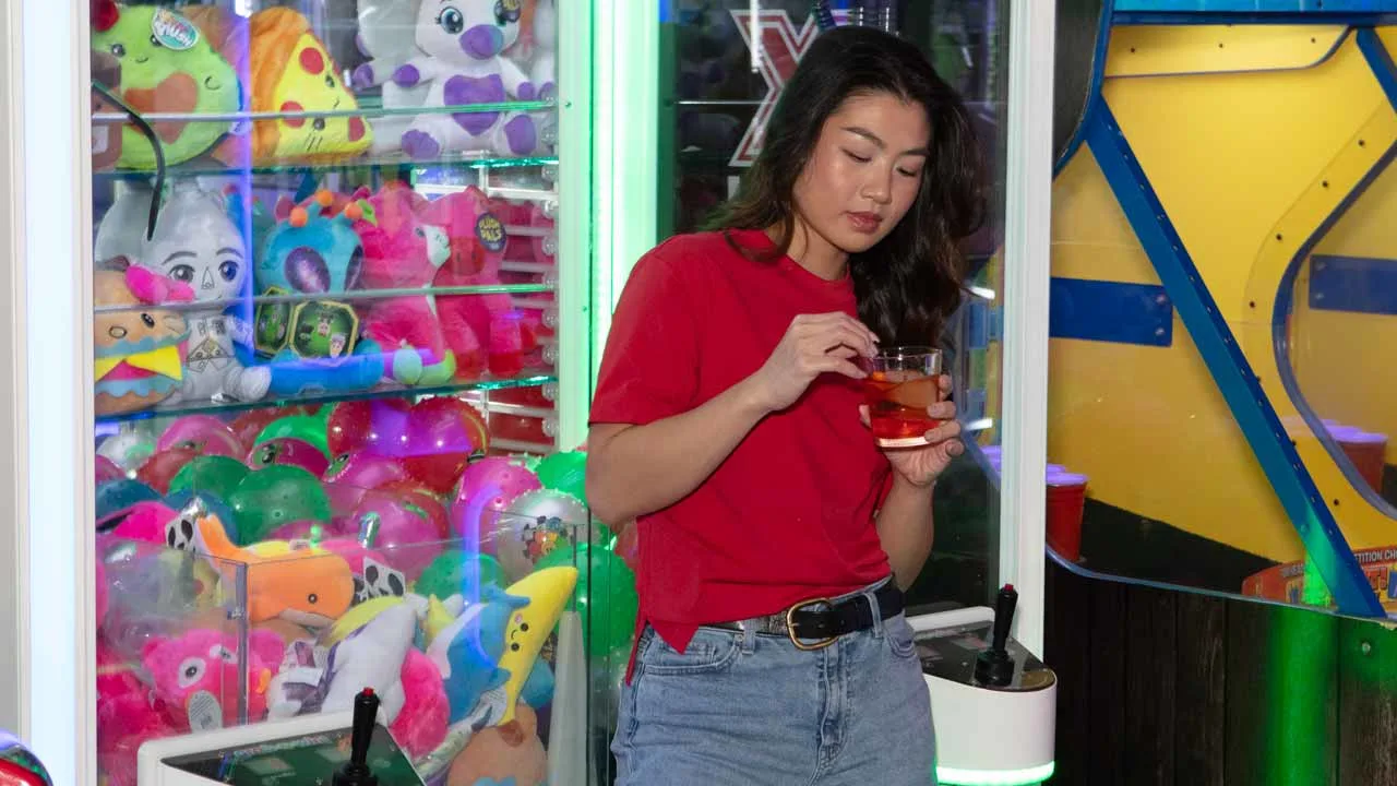A woman holding a drink standing in front of a claw machine.