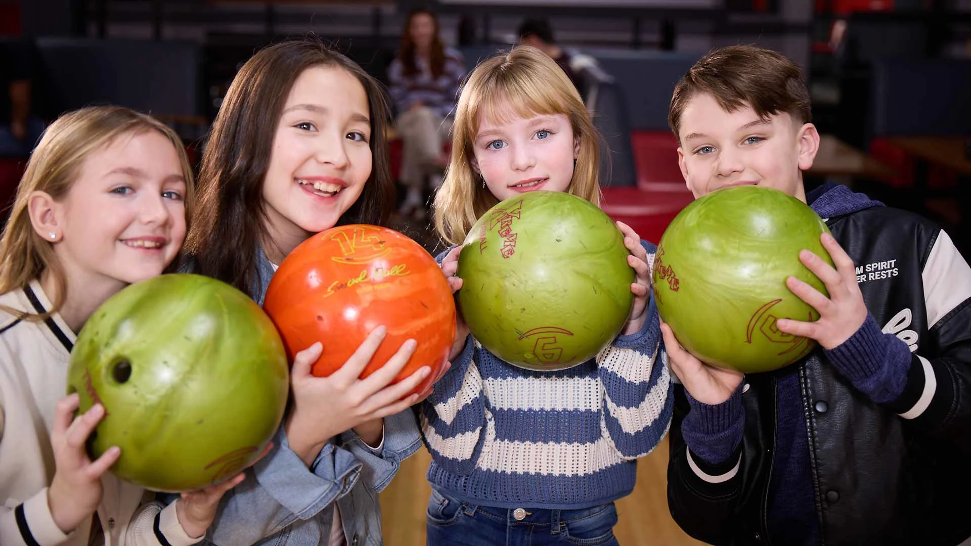 Four smiling kids at a bowling center hold colorful bowling balls and pose together near the seating area.