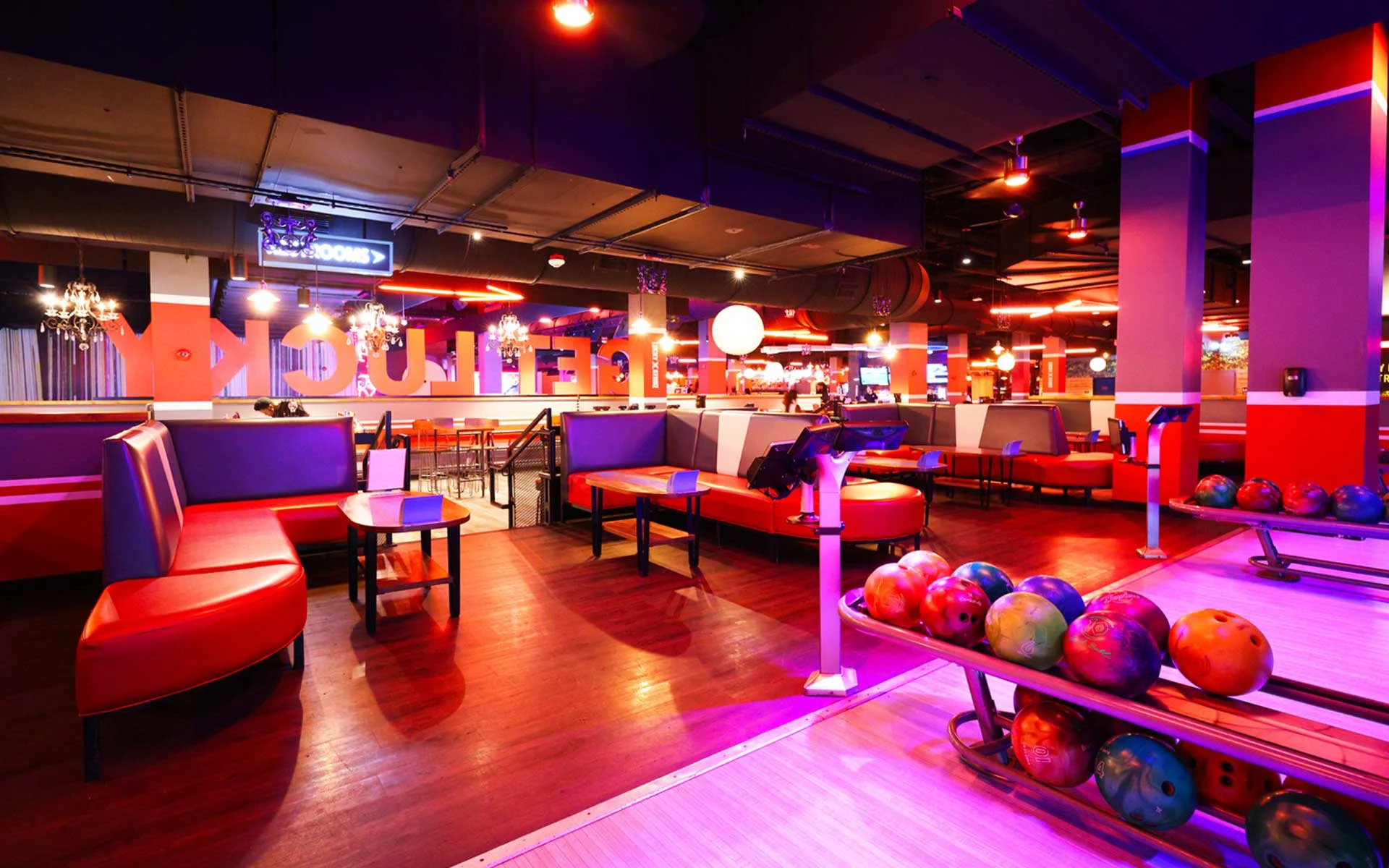 Vibrant bowling lounge with neon lighting, featuring sleek lanes, ball racks, and red booth seating, alongside modern scoring consoles and a lively “Get Lucky” backdrop for an upscale, social atmosphere.