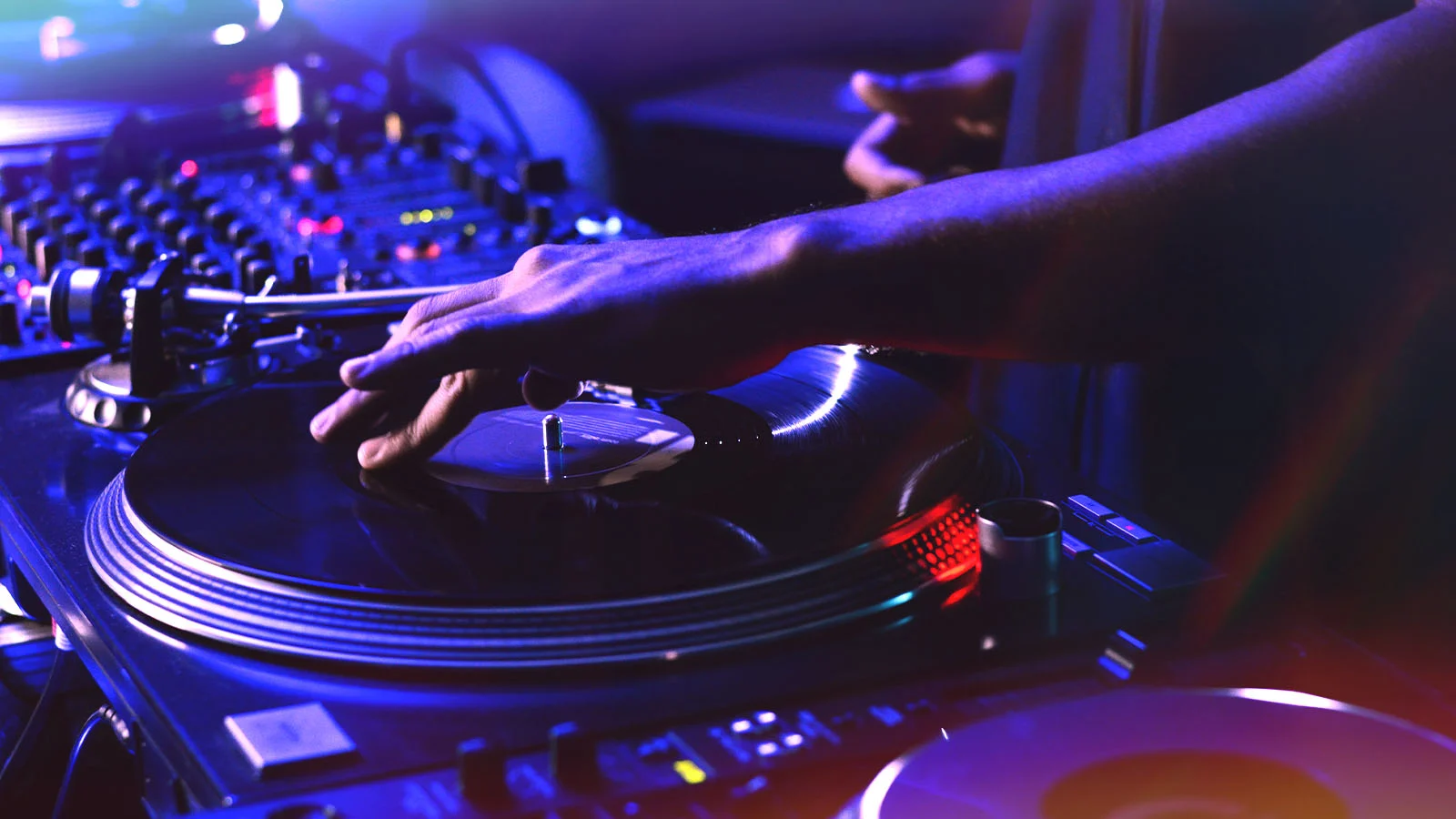 Close-up of a DJ mixing music on a turntable and mixer, creating a live soundtrack with colorful nightlife lighting at an event or party.