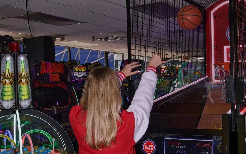 A woman in a red varsity jacket shoots a ball at an arcade basketball hoop game surrounded by AMF arcade machines.
