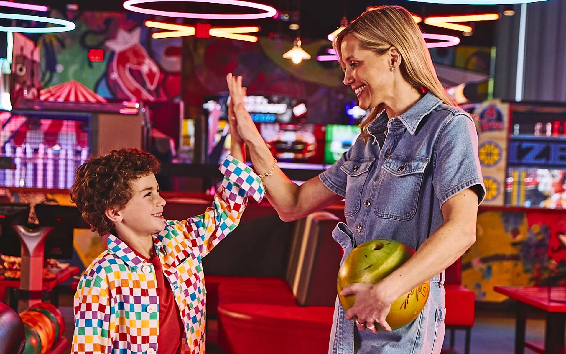Mother and child celebrating with a high-five while holding a bowling ball inside a vibrant, neon-lit bowling and arcade venue, surrounded by colorful seating and games.