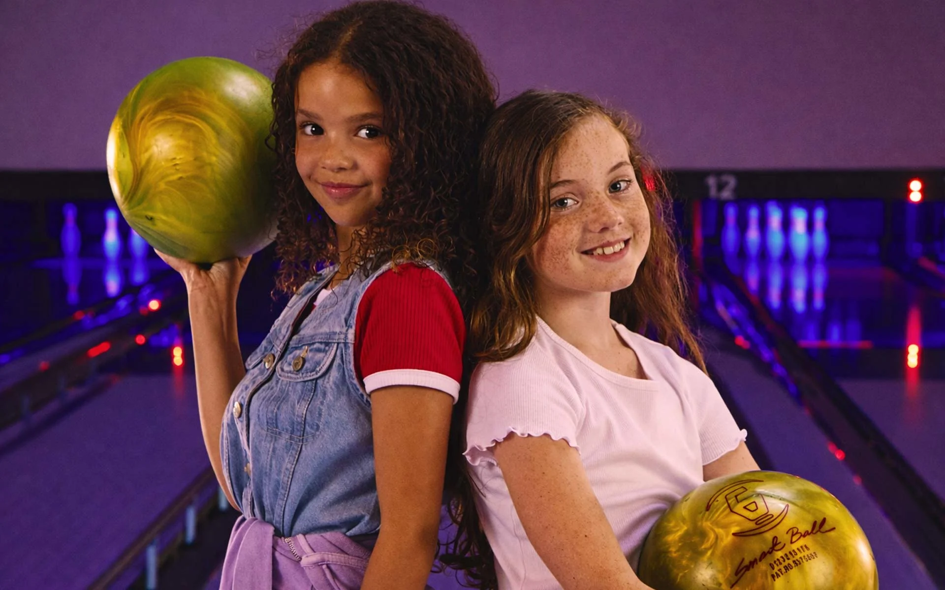 A girl holding a bowling ball and smiling with other kids in the background.