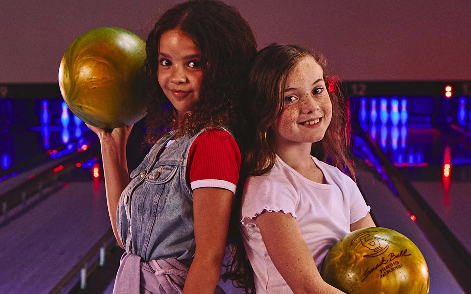 A girl holding a bowling ball and smiling with other kids in the background.