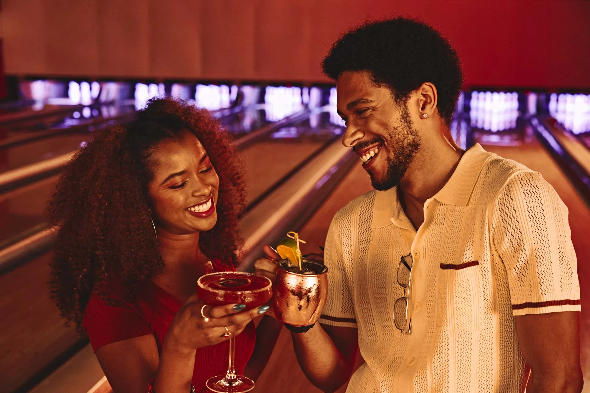 A couple cheering drinks in front of bowling lanes in a Lucky Strike center.