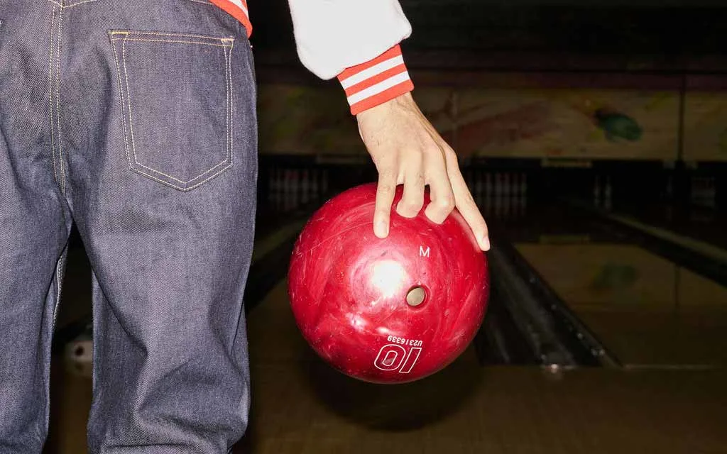 Close-up of a bowler in a varsity jacket and denim holding a red house bowling ball at the top of the lane.
