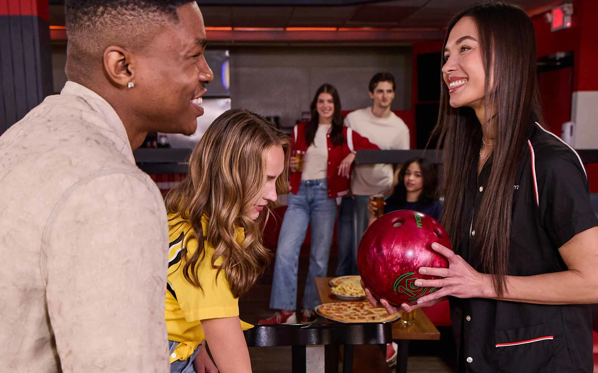Group of friends socializing at a bowling alley, with one woman holding a red bowling ball while others chat and enjoy food and drinks in the background.