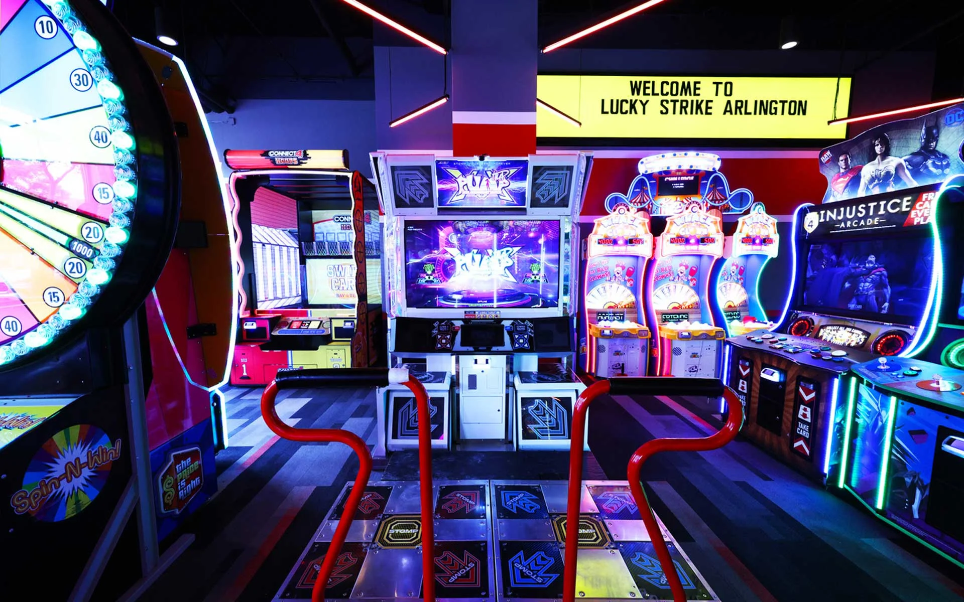 Neon-lit arcade area at Lucky Strike Arlington, featuring a central dance game platform, classic and modern arcade machines, and colorful prize games beneath a bright “Welcome to Lucky Strike Arlington” sign.