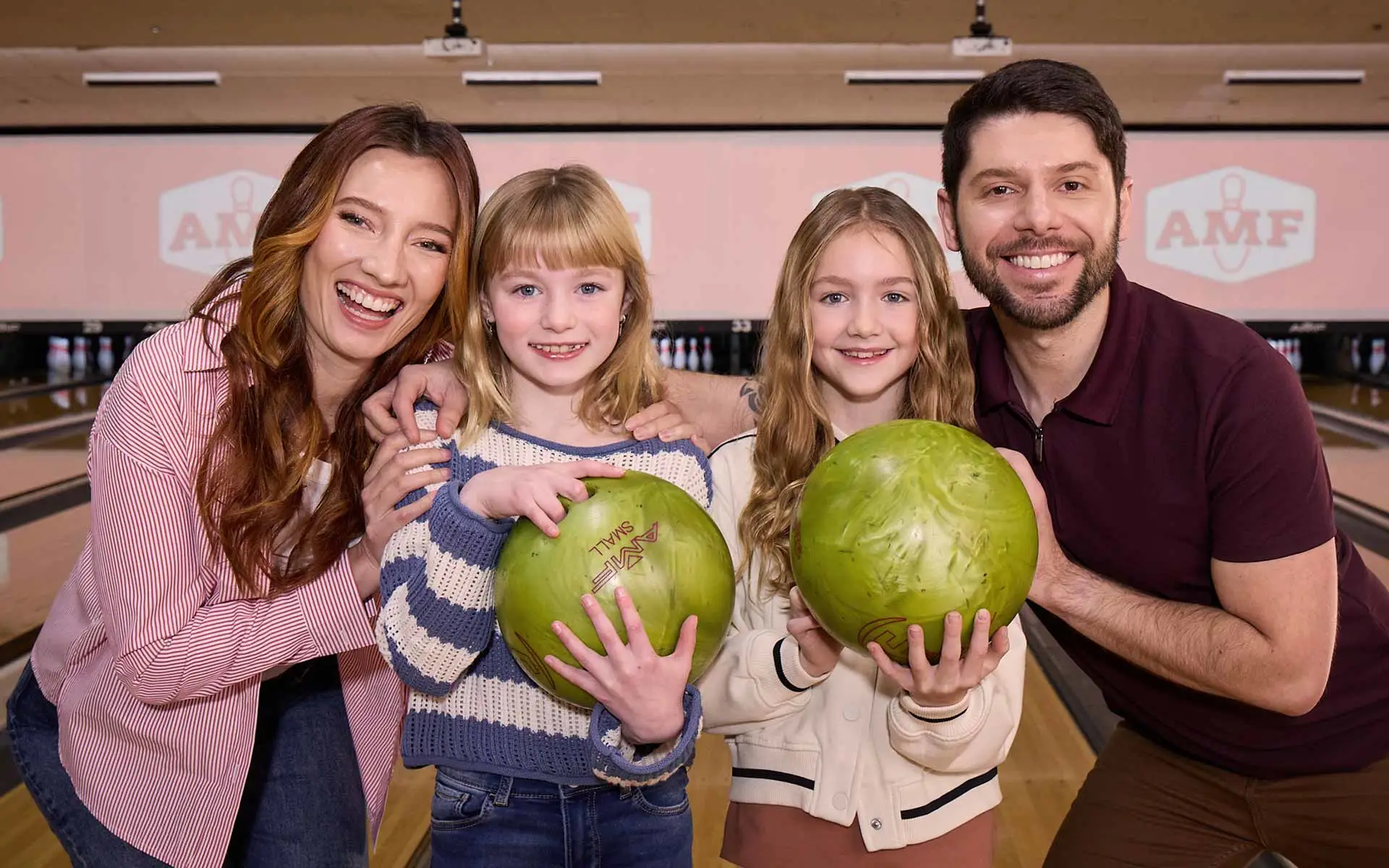 Family of father, mother and two daughters smiling in front of AMF lanes while smiling and having fun bowling.