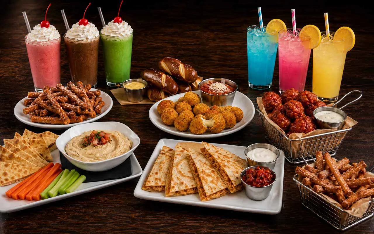 Shareable appetizer spread with pretzels, wings, mozzarella bites, hummus, quesadillas, milkshakes, and colorful lemonades on a wooden table.