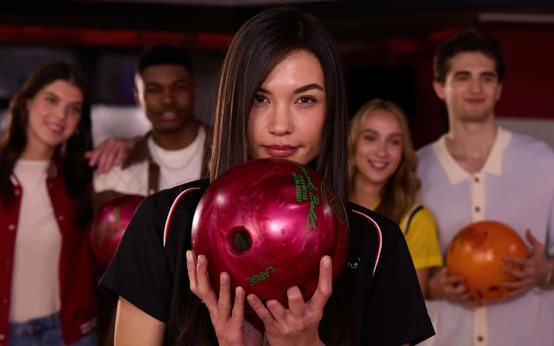 Woman holding a red bowling ball in the foreground while four friends stand behind her with bowling balls inside an AMF bowling center.