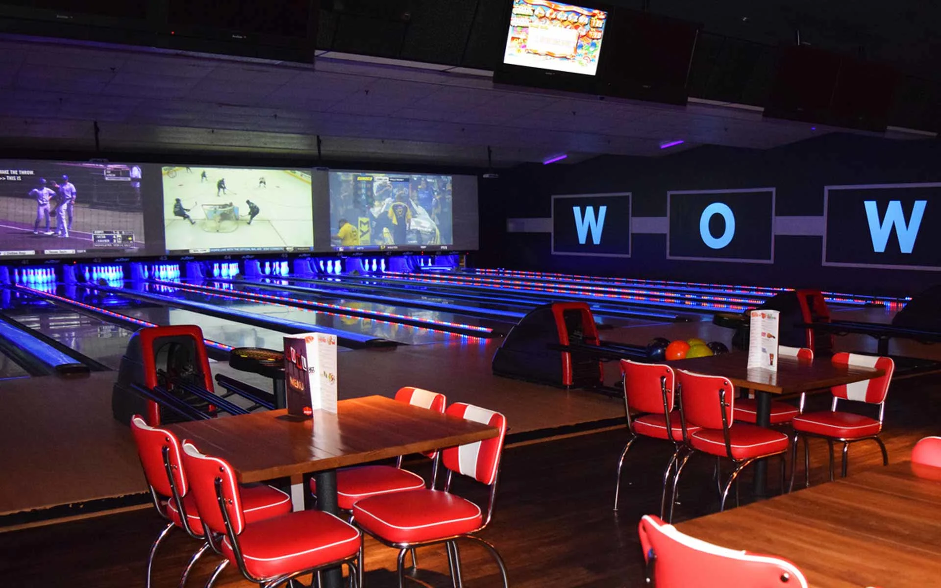 Stylish bowling alley with neon-lit lanes, large projection screens showing live sports, and retro red seating areas with tables, creating a lively, modern entertainment atmosphere.