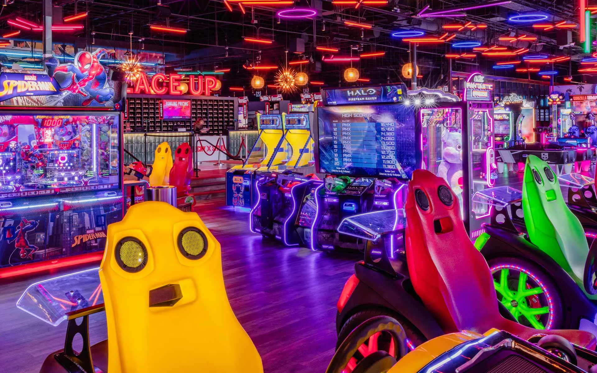 Vibrant arcade floor with neon lighting, featuring racing games, interactive machines, and colorful seating in a high-energy Lucky Strike entertainment venue.