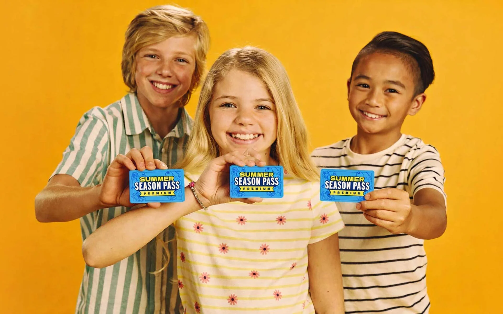 Three smiling kids stand in front of a bright yellow background holding Summer Season Pass Premium cards toward the camera.