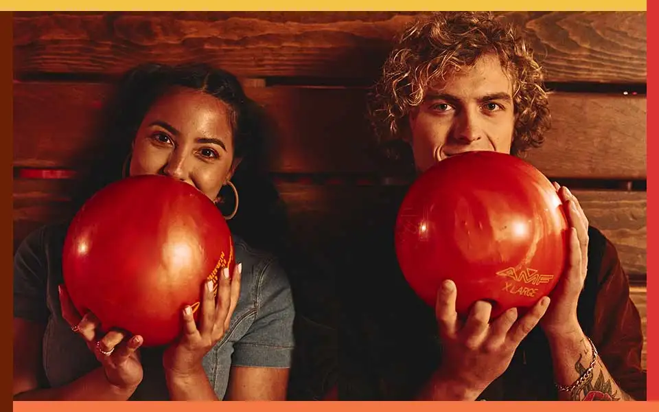 Two bowlers holding bowling balls in front of a wood background
