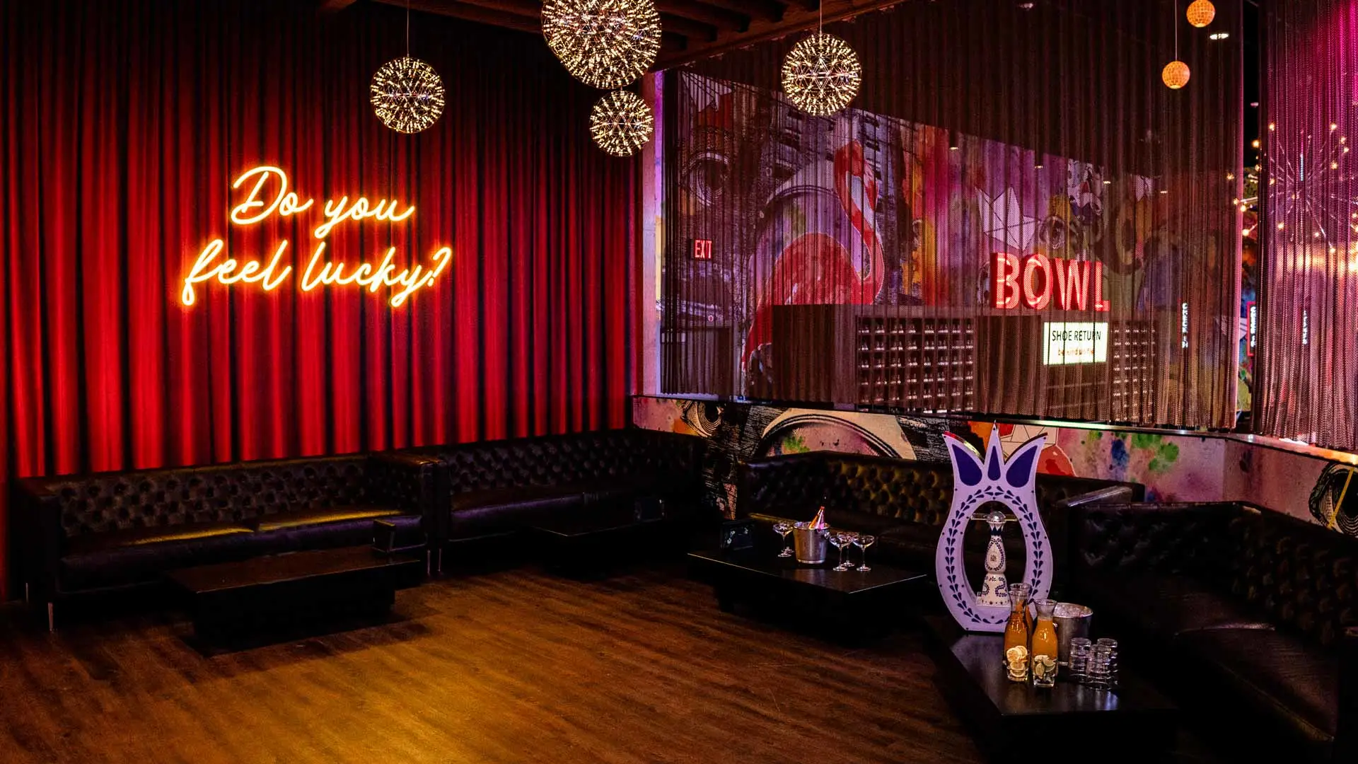 Stylish lounge seating area with neon “Do you feel lucky?” sign, leather couches, and ambient lighting inside a modern bowling entertainment venue.