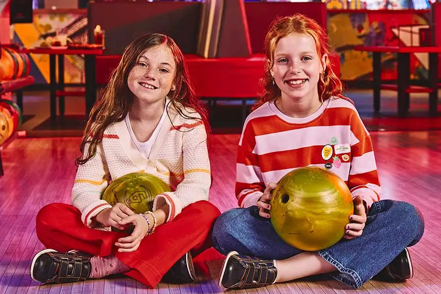 Two young girls sitting on a bowling lane approach holding bowling balls and smiling.