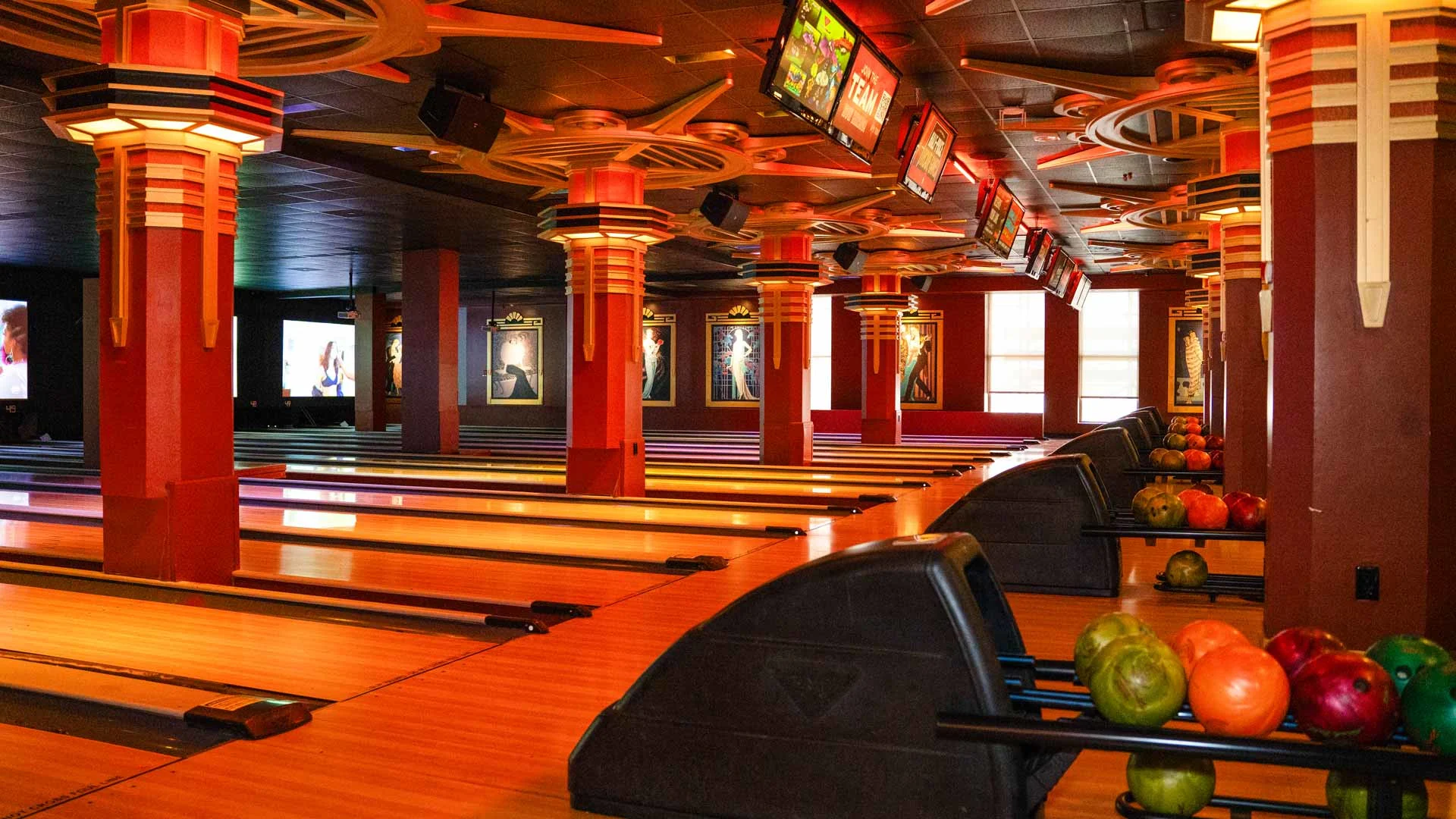 Modern bowling lanes with colorful bowling balls, overhead scoring screens, and art deco–style lighting in an upscale entertainment venue.