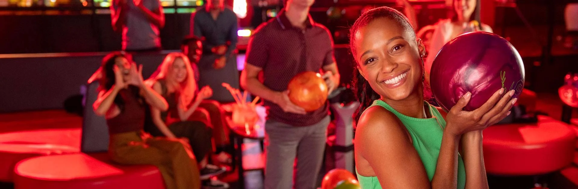 A smiling woman in a green dress holds a purple bowling ball in the foreground of a neon-lit bowling alley while a group of friends sits laughing and cheering on the red couch behind her