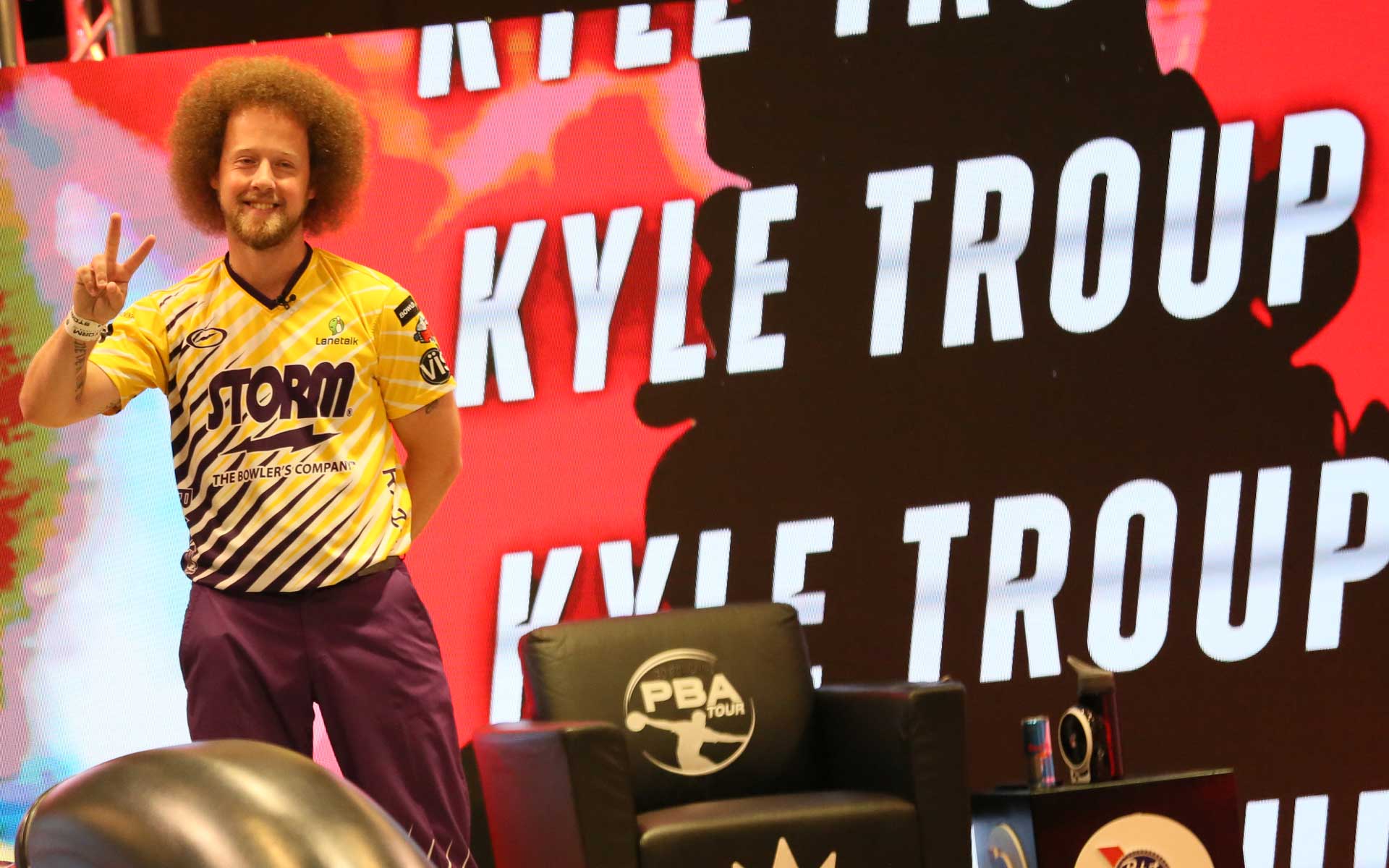 Professional bowler Kyle Troup poses with a peace sign during a PBA Tour event, standing in front of a large illuminated backdrop featuring his name and tour branding.